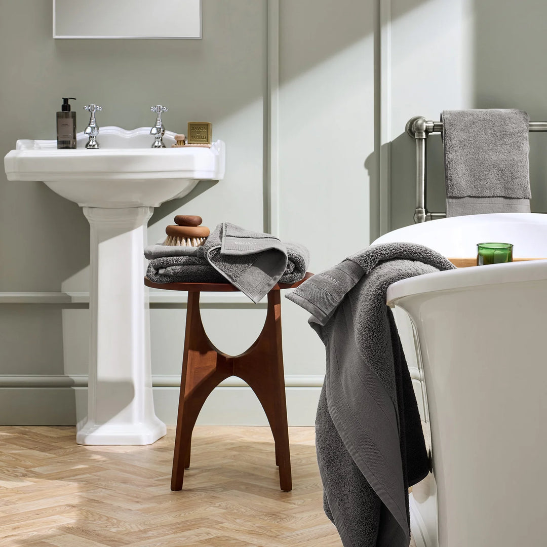 Bathroom with white pedestal sink, wooden stool, and bathtub with gray towels.