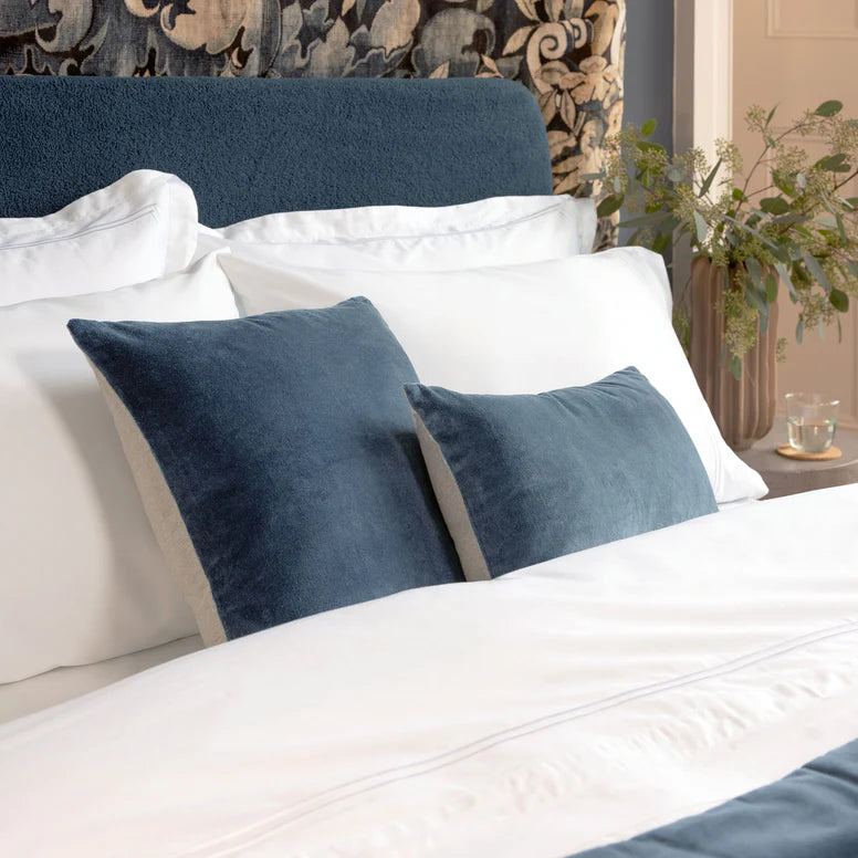 Close-up of a bed with white bedding and blue pillows, featuring a decorative headboard and plant.