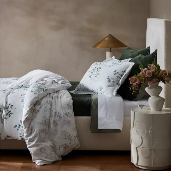 Bedroom with floral bedding and decorative pillows on a bed, next to a side table with a lamp and flowers.