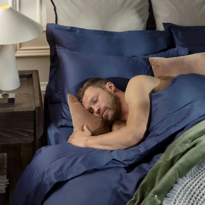 Man sleeping in bed with blue bedding and pillows in a bedroom setting.