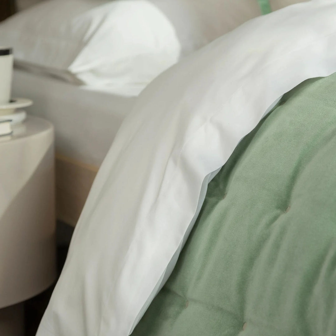 Close-up of a bed with green bedding and white pillows, with a side table in the background.