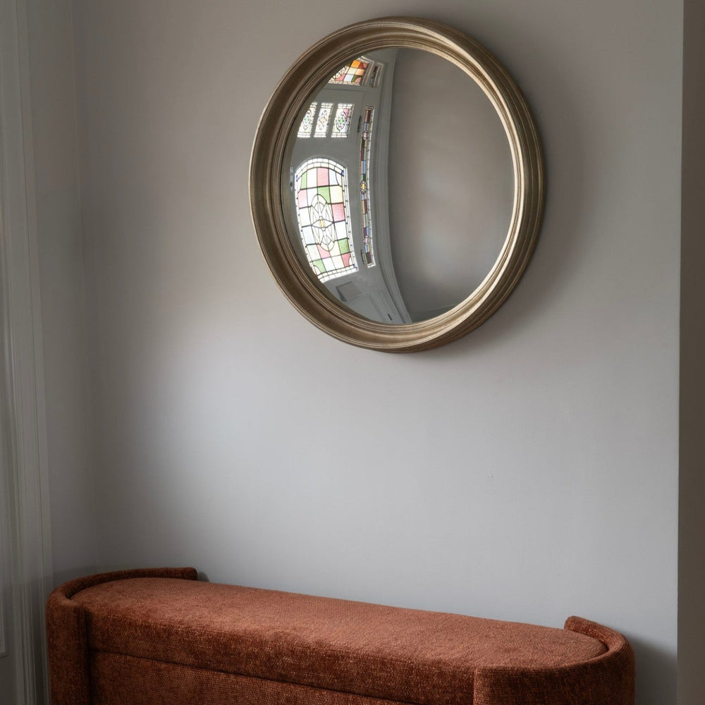 Round mirror with gold frame on a wall above a brown bench with a geometric floor.