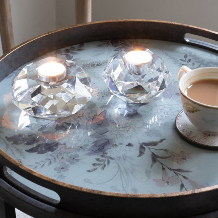 Round wooden tray with crystal candle holders and a cup of tea on a floral-patterned surface.