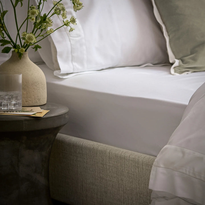 Neatly made bed with white bedding and a small table with a vase and glass of water.
