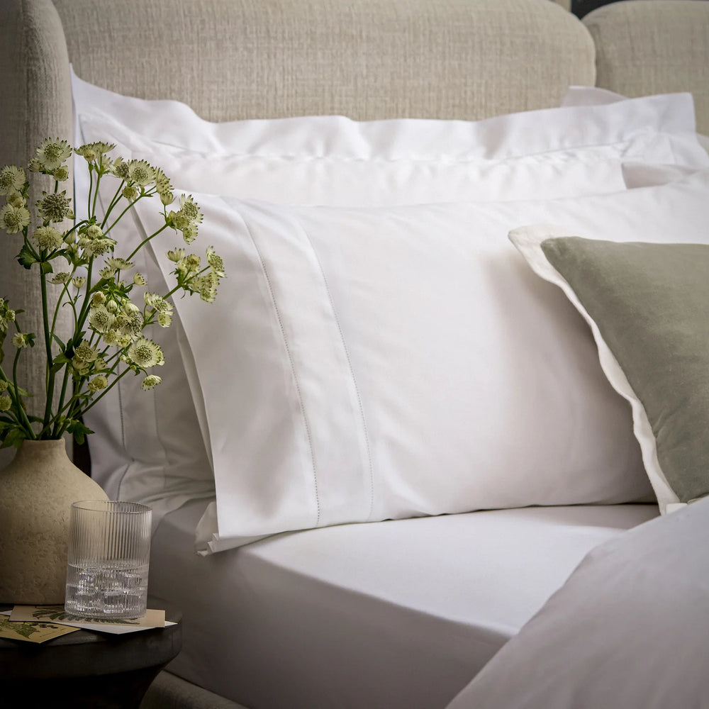 White pillows on a bed with a vase of flowers and a glass on a nightstand.
