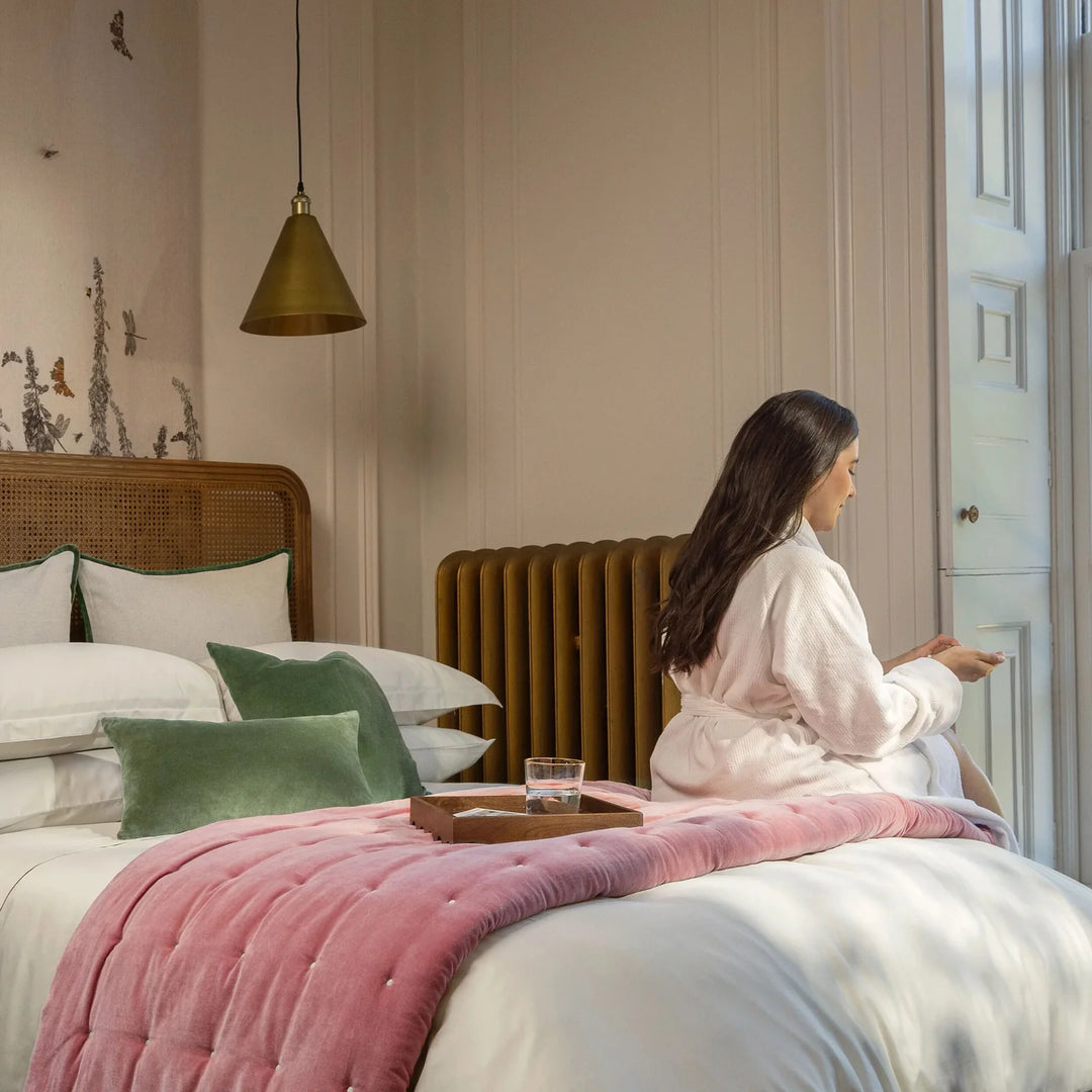 Woman in a robe sitting on a bed in a stylish bedroom with pink blanket and green pillows.