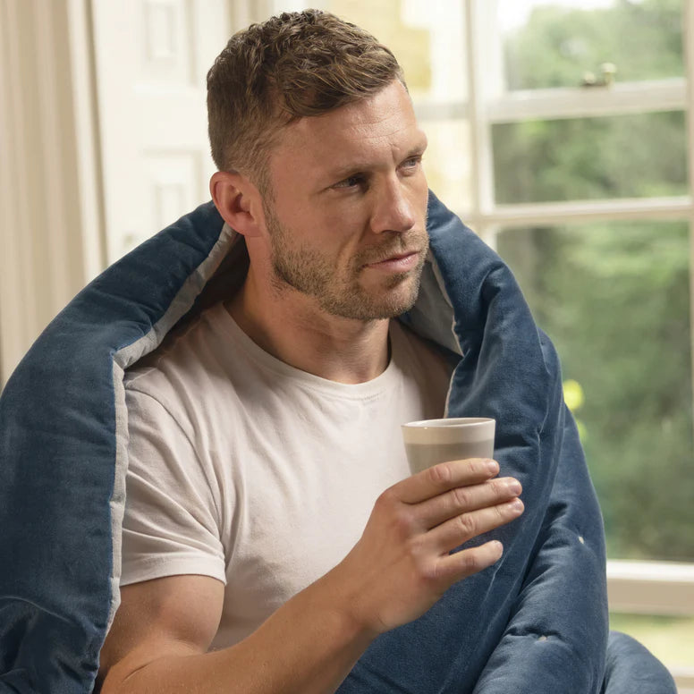 Man wrapped in a blue blanket holding a cup, sitting by a window.