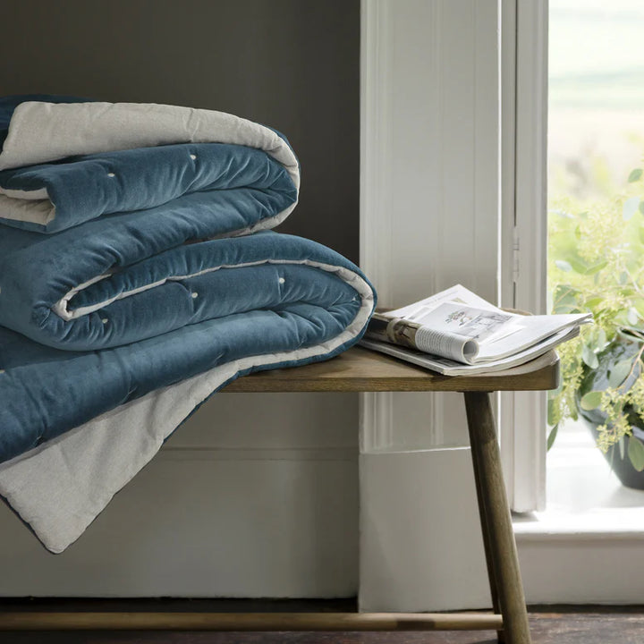 Blue and gray comforter on a wooden bench with a newspaper and plant in a bright room.