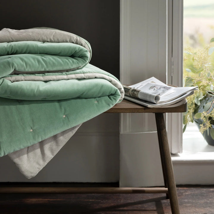 Folded green and gray comforter on a wooden bench by a window with a newspaper.