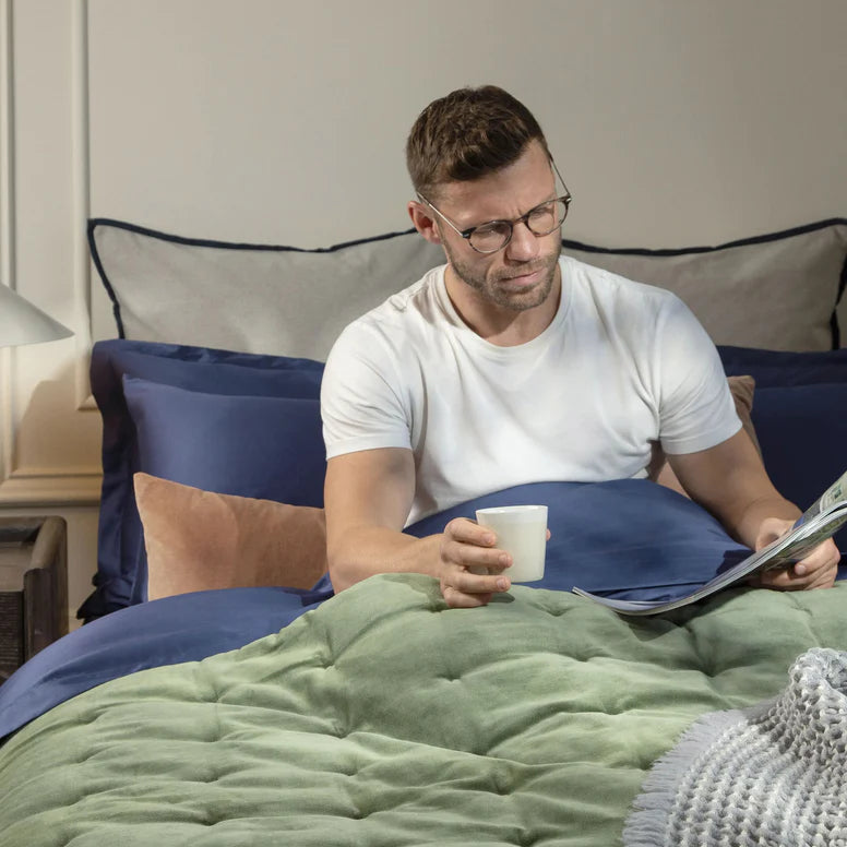 Man sitting on a bed reading a magazine and holding a cup.