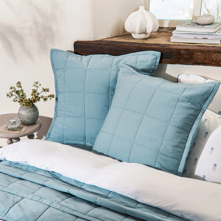 Blue quilted pillows on a bed with a wooden side table and decorative items.