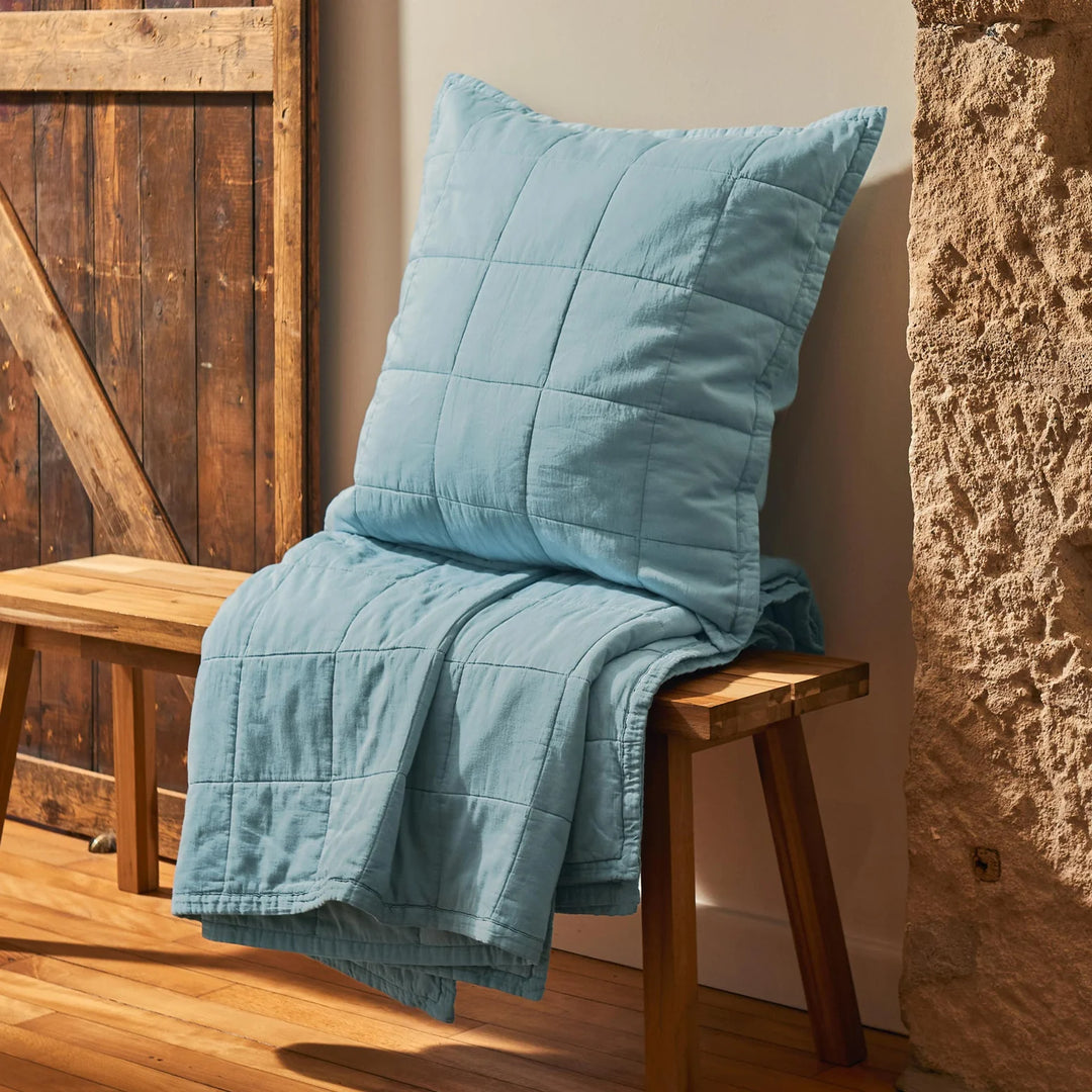 Blue quilt and pillow on a wooden bench against a rustic wall.