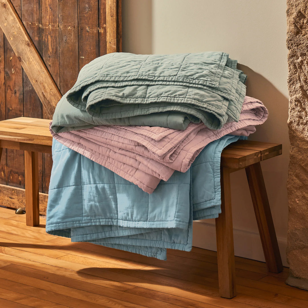 Stack of folded blankets in green, pink, and blue on a wooden bench against a rustic background.