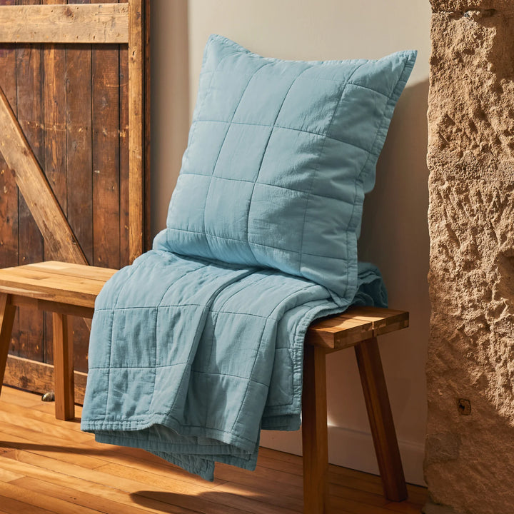 Blue quilt and pillow on a wooden bench against a rustic stone wall.