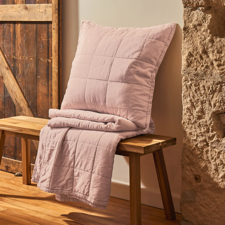 Pink quilt and pillow on a wooden bench against a stone wall.