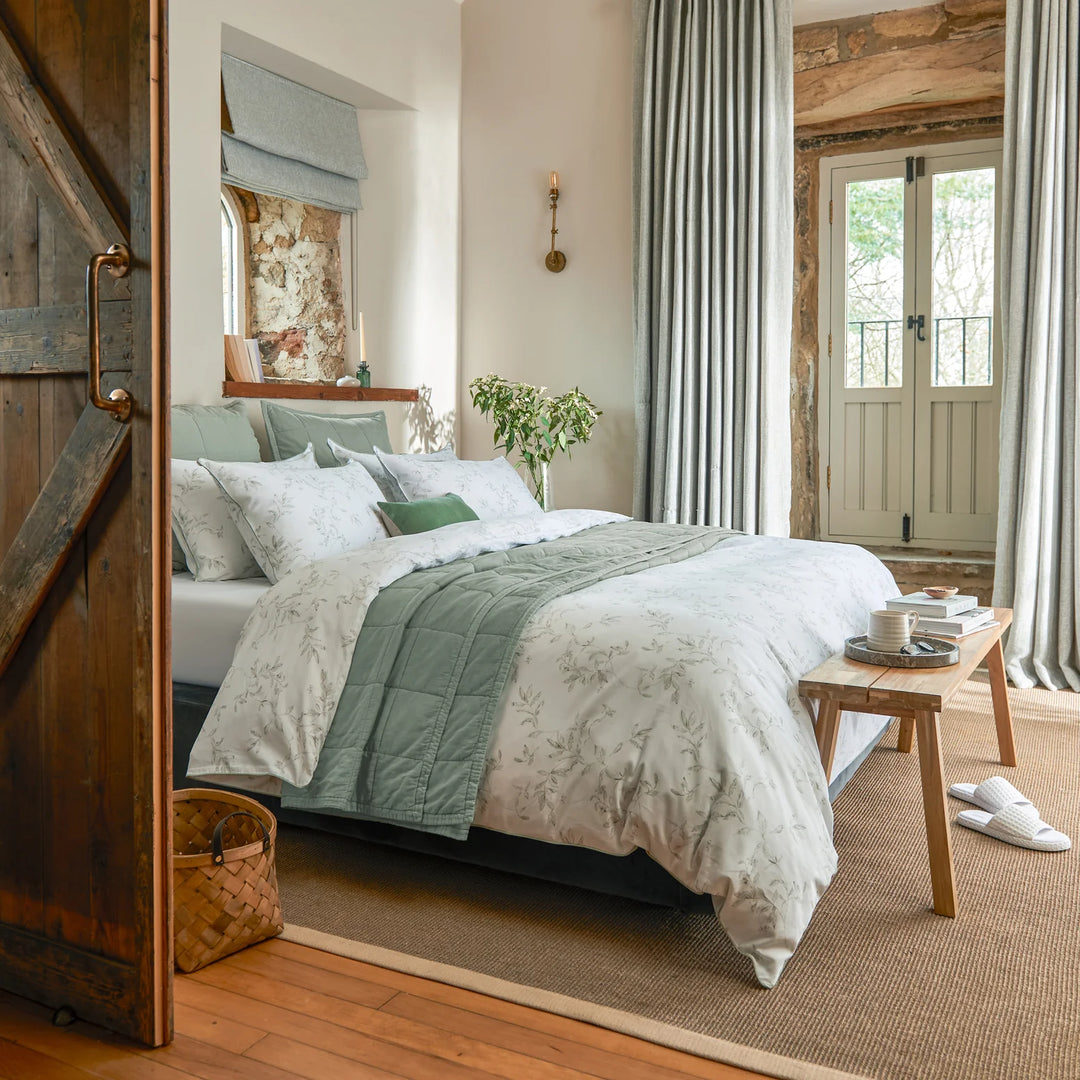 Bedroom with wooden floor, bed with white and green bedding, and a small table with items.