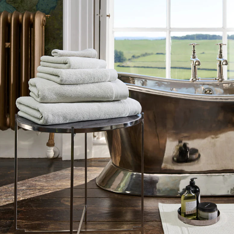 Stack of CHRISTY Luxe - French Grey on a stool next to a freestanding bathtub with a scenic view.