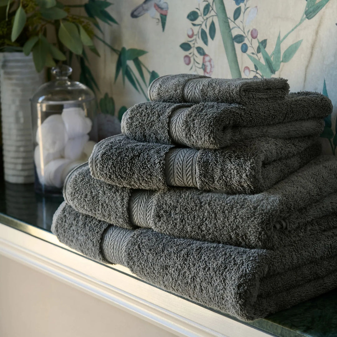 Stack of CHRISTY Renaissance Towels - Ash Grey on a reflective surface with a floral-patterned curtain in the background.