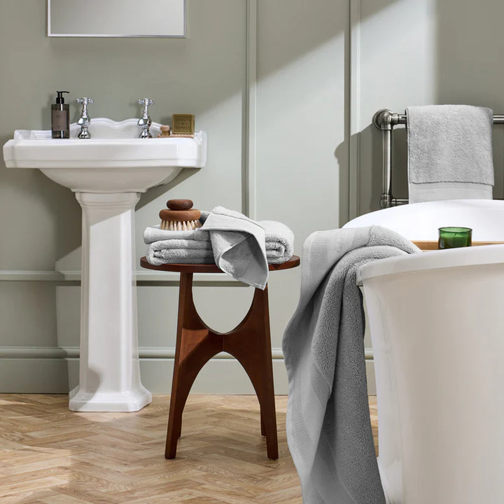Bathroom with white pedestal sink, wooden stool, and bathtub with towels.