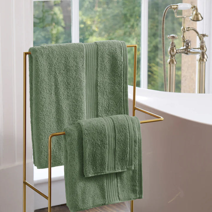 CHRISTY Supreme Hygro Towels - Bay Leaf Green hanging on a gold towel rack in a bathroom with a window in the background.