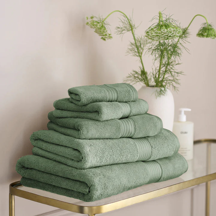 CHRISTY Supreme Hygro Towels - Bay Leaf Green on a gold metal shelf with a vase of greenery in the background.
