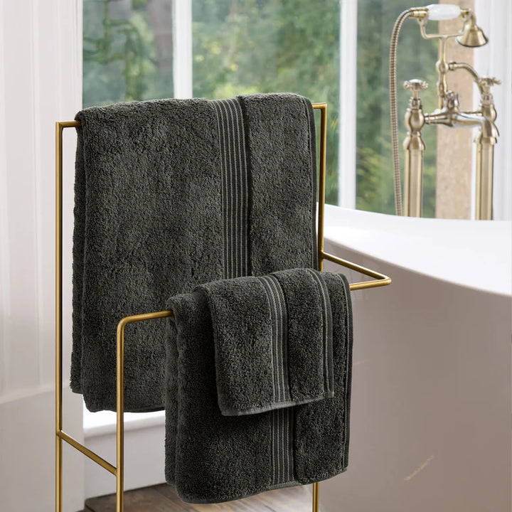 CHRISTY Supreme Hygro Towels - Graphite on a gold towel rack in a bathroom with a bathtub and window in the background.