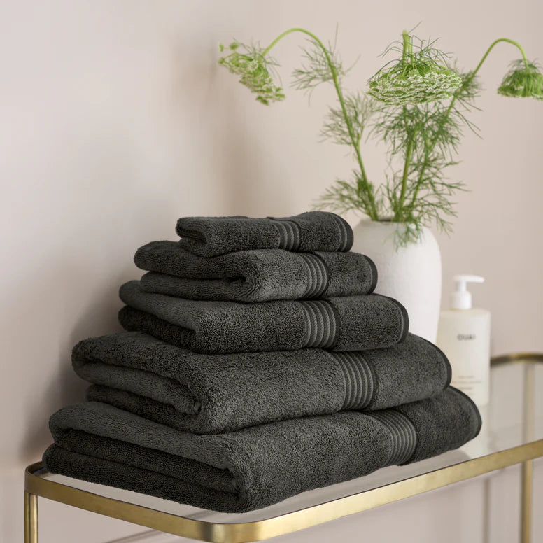 Stack of CHRISTY Supreme Hygro Towels - Graphite on a gold shelf with a vase of greenery in the background.