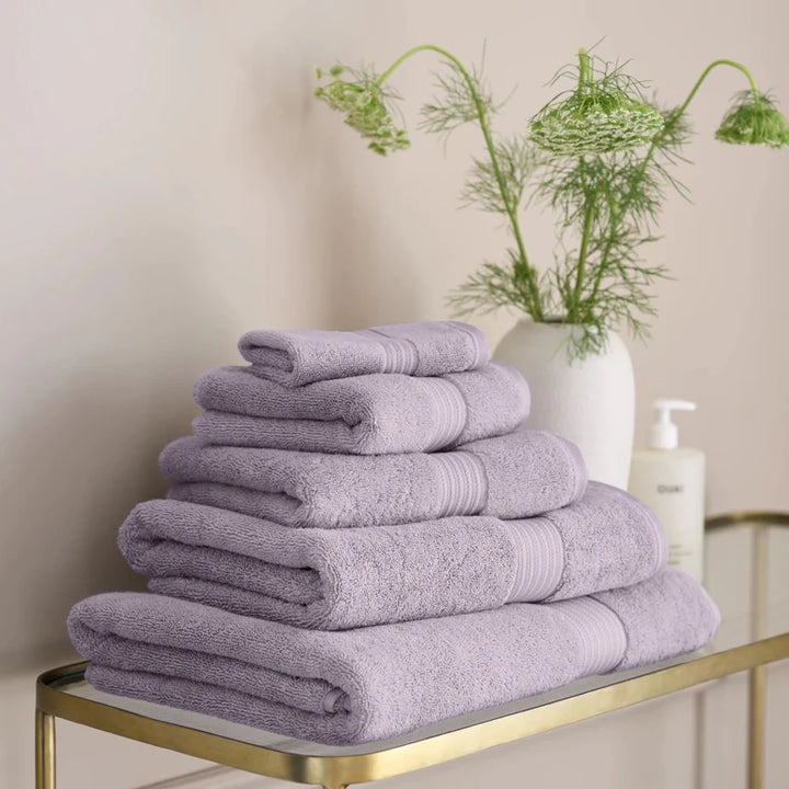 Stack of CHRISTY Supreme Hygro Towels - Lavender on a gold metal shelf with a vase of greenery in the background.