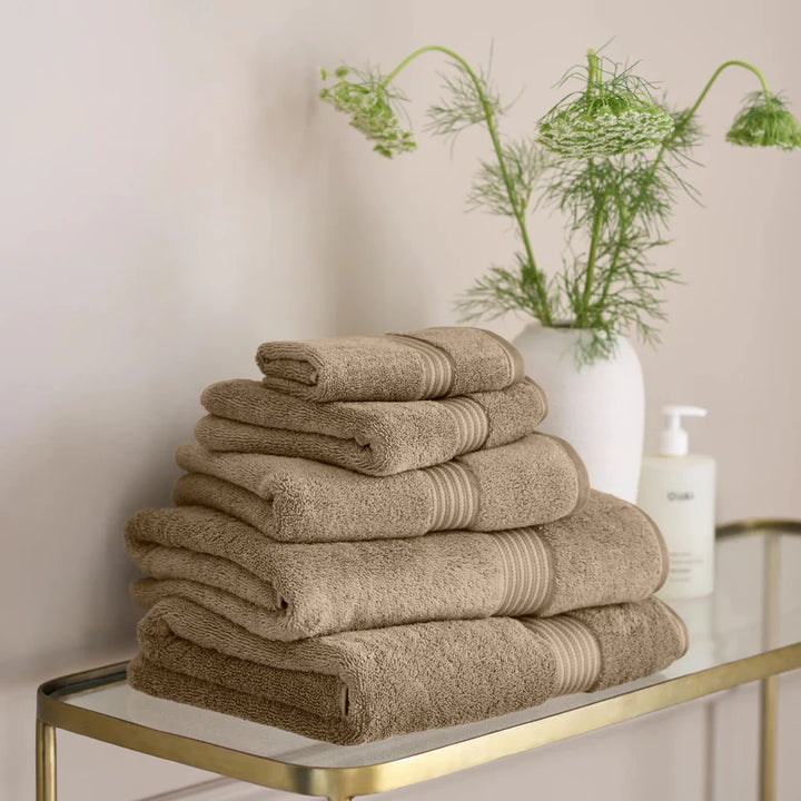 Stack of CHRISTY Supreme Hygro Towels - Mocha on a glass table with a vase of greenery in the background.