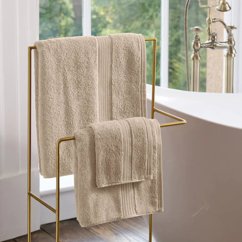 CHRISTY Supreme Hygro Towels - Stone hanging on a gold towel rack in a bathroom with a bathtub and window in the background.