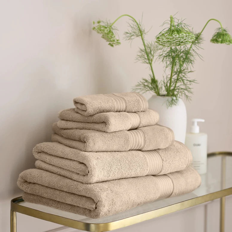 Stack of CHRISTY Supreme Hygro Towels - Stone on a glass table with a vase of green flowers in the background.