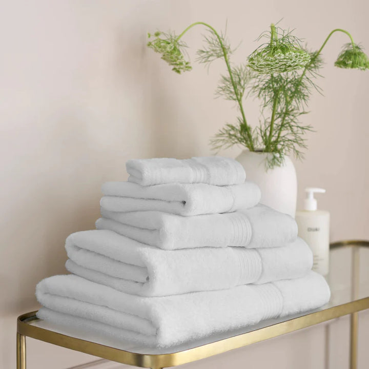 Stack of CHRISTY Supreme Hygro Towels - White on a gold metal shelf with a vase of greenery in the background.