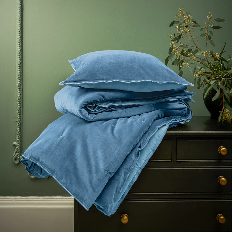 Blue bedding set on a dark dresser against a green wall with a plant.